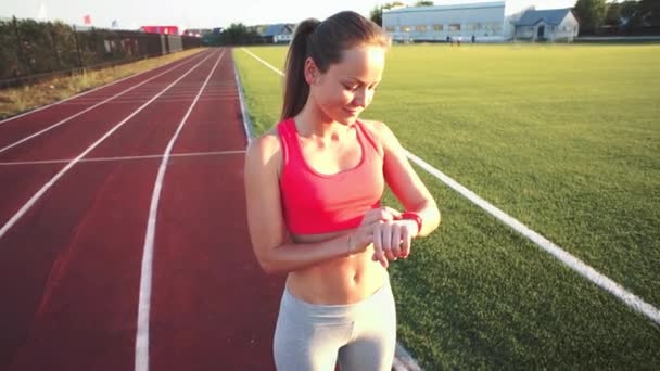 Belle jeune femme configure un programme sur son bracelet et courir sur piste athlétique sur le stade au lever du soleil. Vidéo de style cinématographique . — Video
