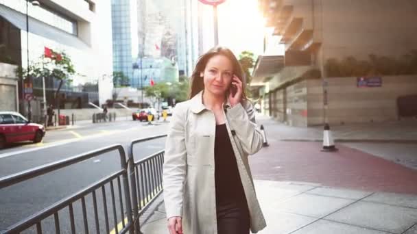 Mujer joven de moda en abrigo blanco ocupado con su teléfono móvil mientras camina por una calle de la ciudad — Vídeos de Stock