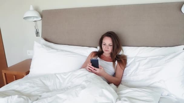 Mujer acostada en la cama revisando mensajes en el teléfono móvil — Vídeos de Stock