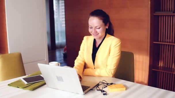 Mulher bonita nova vestida com jaqueta amarela sentada na cadeira do escritório ao usar laptop. Mulher bebendo café e sorrindo . — Vídeo de Stock