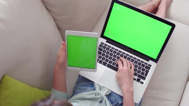 Mooi meisje met tablet pc en laptop met pre-keyed groen scherm zitten op de bank thuis. — Stockvideo