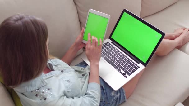 Belle fille en utilisant tablette PC et ordinateur portable avec pré-clavier écran vert assis sur le canapé à la maison. — Video