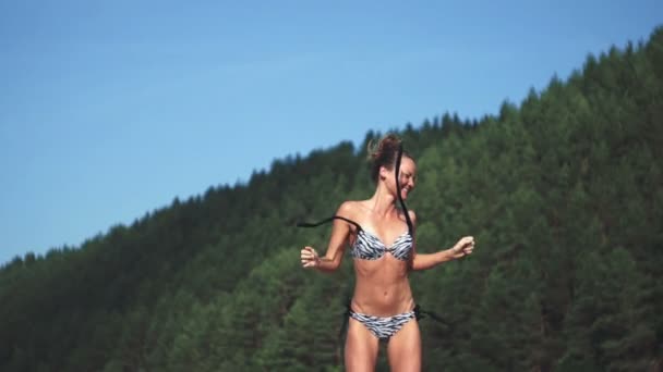 Tiro ao ar livre de sorrir jovem modelo feminino em biquíni dançando e pulando contra o céu azul e floresta verde. Mulher se divertindo em um dia de verão . — Vídeo de Stock