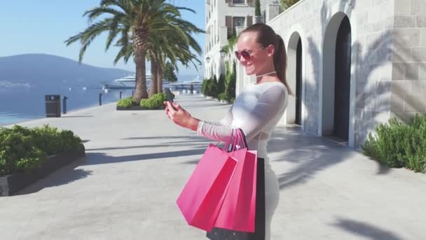 Napszemüvegek a gyönyörű nő a gazdaság bevásárló táskák, használ egy okos telefon, és mosolyogva, miközben állandó szabadban