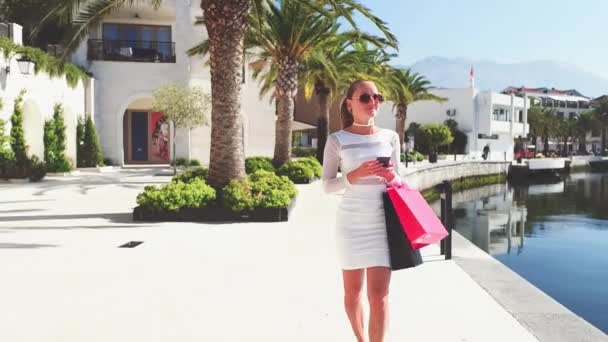 Beautiful woman in sun glasses are holding shopping bags, using a smart phone and smiling while standing outdoors — Stock Video