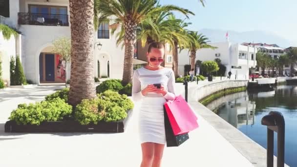 Медленное движение. Красивая женщина в солнечных очках держит сумки для покупок, используя смартфон и улыбаясь стоя на улице — стоковое видео
