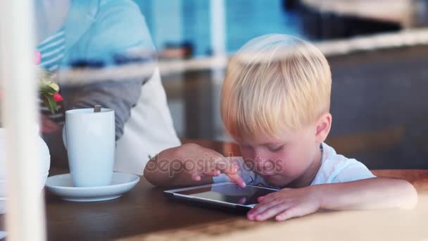 Niño de 4 años sentado en el escritorio de madera mientras juega con el dispositivo de tableta en la cafetería de la ciudad — Vídeos de Stock