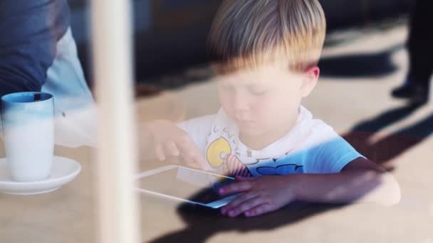 4 years old children boy sitting at the wooden desk while playing with the tablet pad device at the city cafe — Stock Video