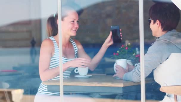 Two young girls smiling using smart phone in a cafe. The photo was taken through the window — Stock Video