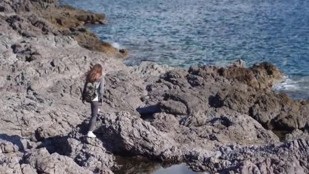 Young woman hiker with backpack standing on cliff and looking forward on the background of the sea, sky. lady tourist on top of a mountain enjoying view. — Stock Video