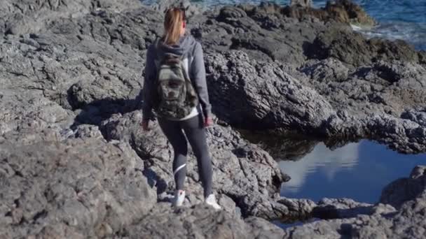 Eine junge Wanderin mit Rucksack steht auf einer Klippe und blickt auf den Hintergrund des Meeres, den Himmel. Touristin auf einem Berg mit Aussicht. — Stockvideo