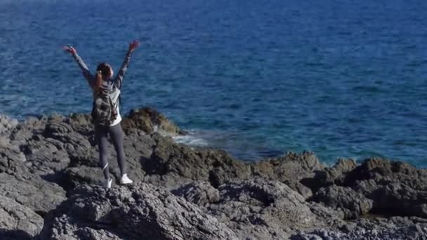 Mujer joven excursionista con mochila de pie en el acantilado y mirando hacia adelante en el fondo del mar, el cielo. mujer turista en la cima de una montaña disfrutando de la vista . — Vídeos de Stock