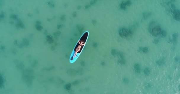 Câmera movendo-se para o topo e em torno de mulher atraente em biquíni é tomar sol em uma prancha de surf, vista aérea — Vídeo de Stock