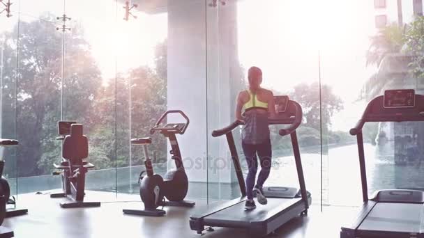 Woman press button and start running on treadmill at a panoramic window and listening to music via headphone at gym. Concept of healthy lifestyle. — Stock Video