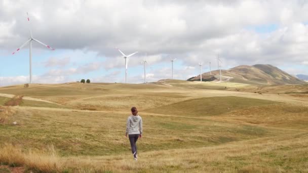 Женщина идет по полю с ветряными турбинами на фоне заката. Производство альтернативной возобновляемой энергии — стоковое видео