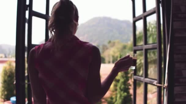 Vista a la montaña. Joven hermosa chica bebe café de la mañana en una ventana en casa en el jardín tropical. — Vídeos de Stock