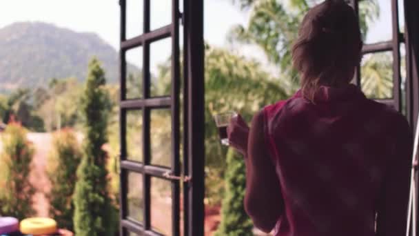 Vista a la montaña. Joven hermosa chica bebe café de la mañana en una ventana en casa en el jardín tropical. — Vídeos de Stock