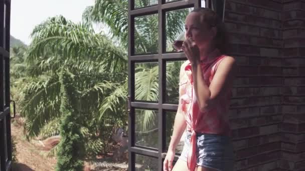 Vista a la montaña. Joven hermosa chica bebe café de la mañana en una ventana en casa en el jardín tropical. — Vídeos de Stock