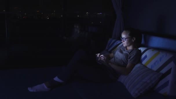 Woman using the laptop and sitting beside the window on her bad. Tv light. — Stock Video