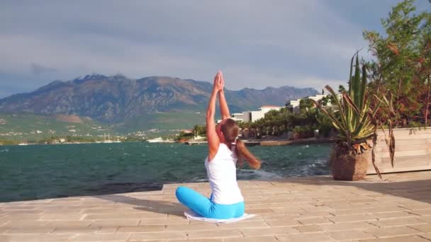 Jeune femme asiatique en pose de yoga à la jetée de villa privée. Femme sportive vêtue de blu et de vêtements blancs faisant du yoga avec vue sur la montagne à la mer adriatique au Monténégro . — Video