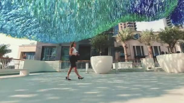 Joven mujer de negocios sonriente en vasos y taza de café caminando por la ciudad con edificios modernos en el fondo. — Vídeo de stock
