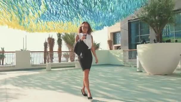 Giovane donna d'affari sorridente in occhiali e tazza di caffè che cammina in città con edifici moderni sullo sfondo. — Video Stock