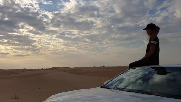 Jonge vrouw zit op het dak van een auto en kijkt naar de epische zonsondergang, tegen de achtergrond van de woestijn. — Stockvideo