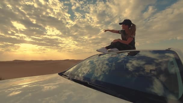 Young woman sitting on the roof of a car and shows by her hfinger on the epic sunset, against the background of the desert. — 비디오