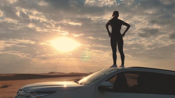 Jovem mulher despreocupada vestida casual desfrutando de paisagem arenosa deserto de pé no telhado do carro com as mãos levantadas no deserto dos Emirados Árabes Unidos — Vídeo de Stock