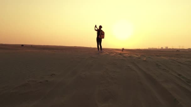 Back view blond hair woman with yellow authentic backpack looking on sandy road after sandstorm. Travel adventures in desert. — Stock Video