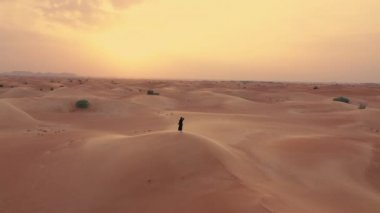 Havadan. Geleneksel Emirati elbisesi içinde genç ve güzel beyaz bir kadının durağan manzarası - çöldeki abaya güçlü rüzgar ve turuncu susnet..