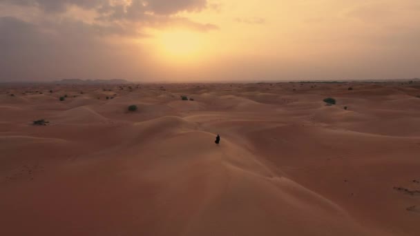 AERIAL. Kamera folgt einer Frau in traditioneller Kleidung der Emirate, die in einer Wüste bei starkem Wind und Sonnenuntergang spaziert. — Stockvideo