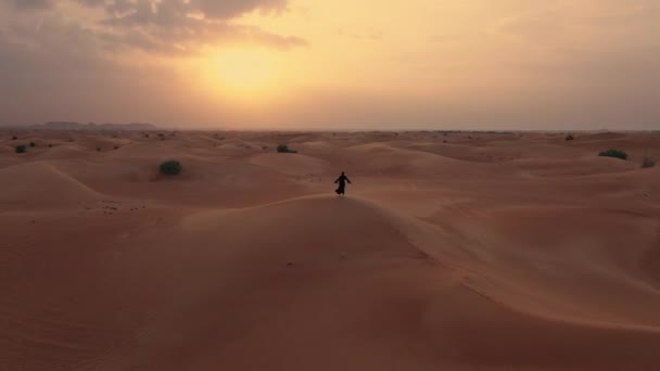 AERIAL. Cámara que se mueve alrededor de la mujer en vestido tradicional emiratí caminando en un desierto en el viento del strog y puesta de sol . — Vídeos de Stock