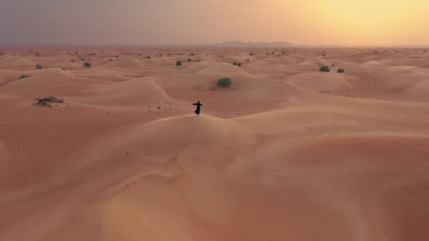 AERIAL. Cámara que se mueve alrededor de la mujer en vestido tradicional emiratí caminando en un desierto en el viento del strog y puesta de sol . — Vídeos de Stock