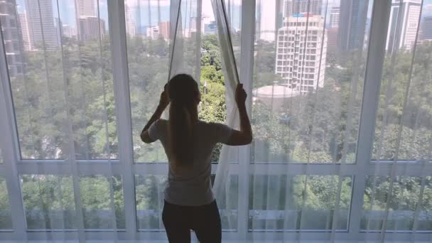 Mujer abre cortina en las grandes ventanas vista de la ciudad con gadern verde y edificios. Concepto de ciudad verde. — Vídeos de Stock