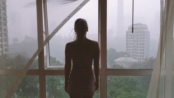 Вид сзади женщины, смотрящей в окно на город под дождем. — стоковое видео