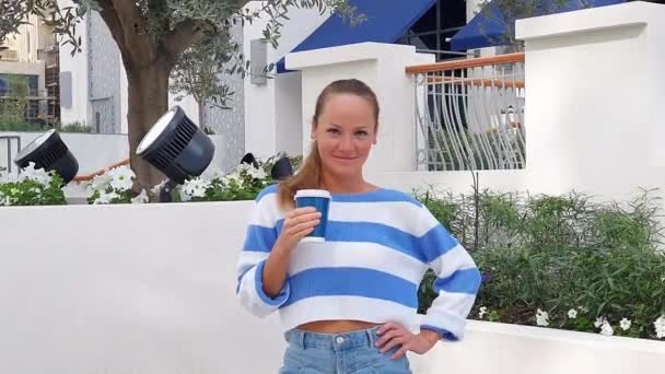 Közelkép egy fiatal nőről, aki kék-fehér kabátban volt. Egy hölgy kávét iszik tavasszal.. — Stock videók