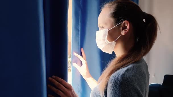 Une femme à la maison regarde par la fenêtre dans un masque médical, mise en quarantaine en raison de la propagation du coronavirus — Video