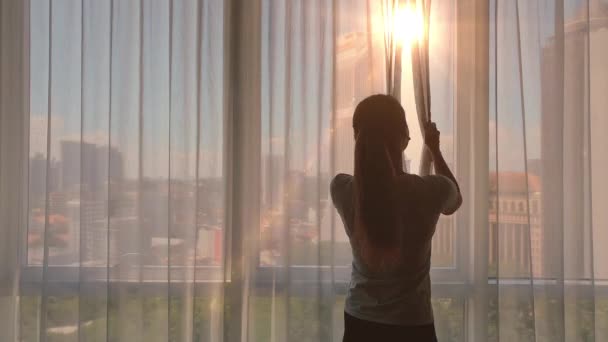 Langzame beweging. Jonge vrouw ontdekt het grote raam en kijkt uit over haar appartement op de gebouwen van de stad. Zonsopgang in de stad. — Stockvideo