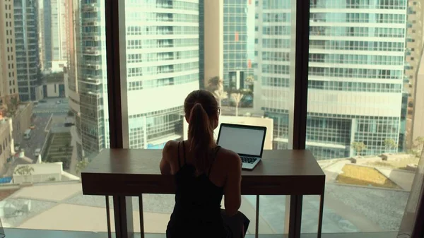 Self-quarantined woman with laptop working near window with city view.