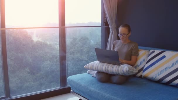 Una joven que trabaja en casa. Freelancer usando laptop, teléfono e Internet. Lugar de trabajo en la sala de estar en alféizar de la ventana. Hora de la cuarentena. COVID - Concepto 19. — Vídeo de stock