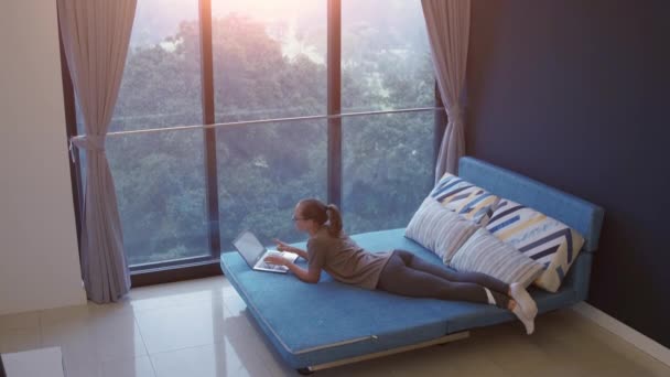 Una joven que trabaja en casa. Freelancer usando laptop, teléfono e Internet. Lugar de trabajo en la sala de estar en alféizar de la ventana. Hora de la cuarentena. COVID - Concepto 19. — Vídeos de Stock