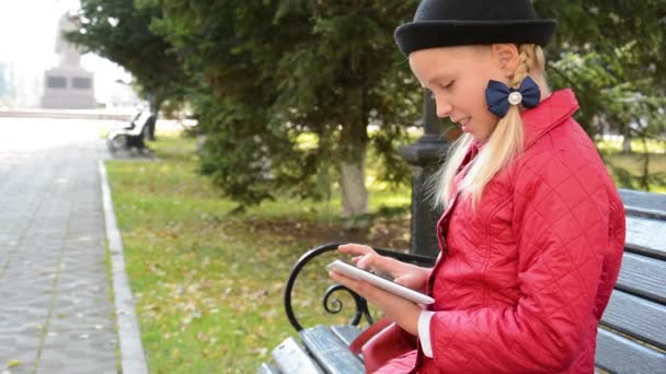 Девушка с планшетным компьютером осенью сидит на скамейке запасных в городском парке. Девушка играет в игру на своем планшетном компьютере — стоковое видео