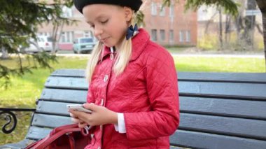 Akıllı telefonda uygulama kullanan genç bir kız sonbaharda şehir parkında bankta oturur.