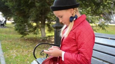 Akıllı telefonda uygulama kullanan genç bir kız sonbaharda şehir parkında bankta oturur.