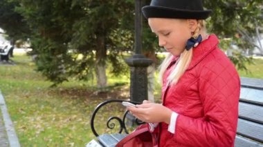 Akıllı telefonda uygulama kullanan genç bir kız sonbaharda şehir parkında bankta oturur.