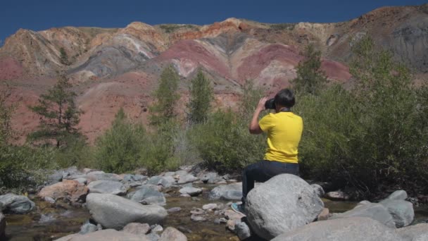 Uma mulher viajante senta-se em uma rocha no meio de um rio de montanha e tira fotos da paisagem — Vídeo de Stock