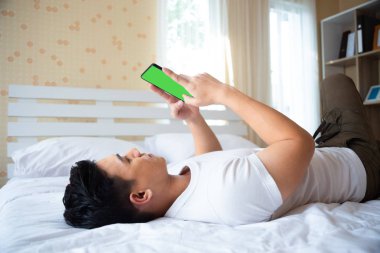 Yakışıklı genç adam yatakta uzanıyor ve akıllı telefon kullanıyor.