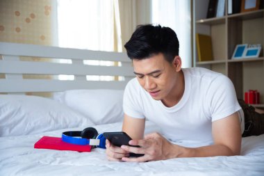 Yakışıklı genç adam yatakta uzanıyor ve akıllı telefon kullanıyor.