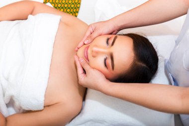 Yatakta ve masözde uzanan genç bir kadın, kan dolaşımı ve sağlık için güzellik merkezinde genç bir bayanla yüz masajı yapmak için iki elini kullanır.